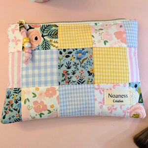 Peut inclure: Une pochette en tissu rectangulaire avec un motif patchwork composé de vichy jaune, de vichy bleu clair et de motifs floraux roses, bleus et verts. Une étiquette indique Noaness Création. La tirette de fermeture éclair a une breloque perlée.