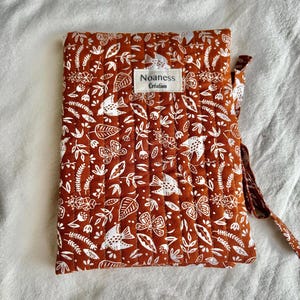 Peut inclure: Une housse en tissu matelassé rectangulaire de couleur orange brûlé avec un motif blanc de plantes et d'oiseaux. Une étiquette rectangulaire blanche est centrée sur le devant avec le texte noir Noaness Création. Un lien en tissu dépasse sur le côté droit.