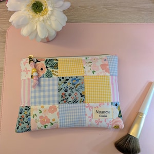 Peut inclure: Une pochette zippée en patchwork avec des motifs floraux, vichy et rayés aux couleurs pastel. La pochette a une fermeture éclair dorée et un charm perlé. Un pinceau de maquillage repose sur une surface rose.