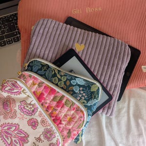 Puede incluir: Una colección de estuches y fundas con cremallera que incluye una funda grande de pana coral con el texto "Girl Boss", un estuche de pana lavanda con un corazón dorado, un estuche floral verde azulado y un estuche crema con estampado de cachemira rosa y morado.