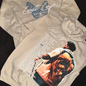 May include: A tan hoodie with a blue butterfly graphic and the text "Last Lap Rod Wave" on the front. The back of the hoodie features a large image of a person performing on stage with a microphone, and a list of tour dates.