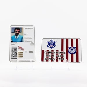 Puede incluir: Dos artículos sobre soportes transparentes. Uno es una tarjeta blanca con una foto, el texto "CYBER, JEFF" y un gráfico de la bandera estadounidense. El otro es una placa roja, blanca y azul con el texto "DOING MORE WITH LESS SINCE 1790" y un emblema de la Guardia Costera.