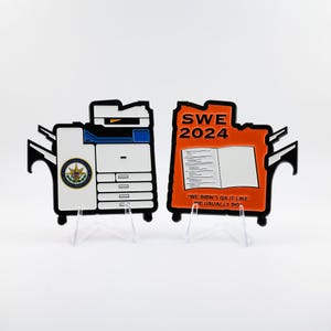 May include: Two novelty items shaped like a copier and a document. The copier is white with black accents and a circular emblem. The document is orange with the text "SWE 2024" and "WE DIDN'T QA IT LIKE WE USUALLY DO."
