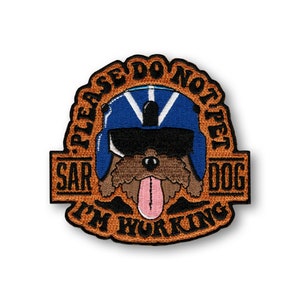 May include: A brown embroidered patch with a cartoon dog wearing a blue helmet and sunglasses. The text on the patch reads "Please Do Not Pet SAR Dog I'm Working".