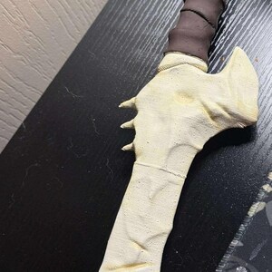 May include: A handcrafted, off-white dagger with a bone-like appearance. The handle is wrapped in dark brown material. The blade features three small, pointed protrusions. The dagger is likely a decorative or cosplay item.