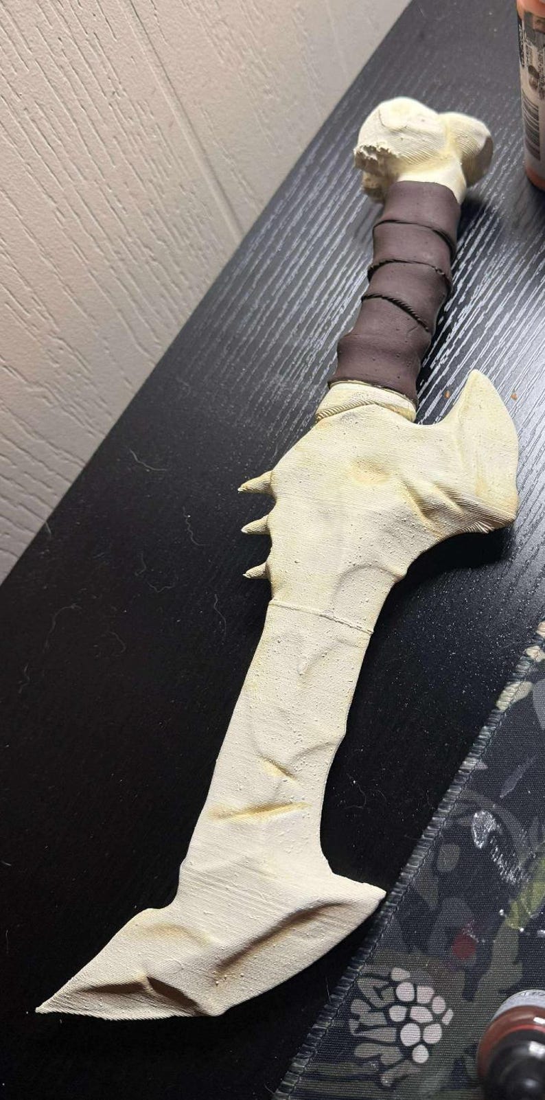 May include: A handcrafted, off-white dagger with a bone-like appearance. The handle is wrapped in dark brown material. The blade features three small, pointed protrusions. The dagger is likely a decorative or cosplay item.