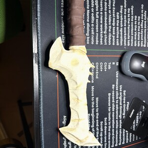 May include: A bone-colored, fantasy-style dagger with a brown wrapped handle. The blade has a textured surface and a stylized design. The dagger is lying on a black surface with computer-related text and a mouse.