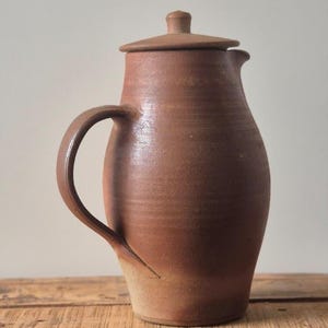 Puede incluir: Una jarra de cerámica marrón con tapa y asa curva. La jarra tiene un pico estrecho y un cuerpo redondeado. La tapa tiene un pequeño pomo en la parte superior. La jarra está sobre una superficie de madera.