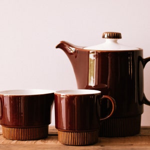 Puede incluir: Un juego de café de cerámica marrón con una tetera con tapa blanca y dos tazas a juego. El juego tiene un diseño retro con una base acanalada y un acabado brillante. Las tazas y la tetera tienen un exterior marrón e interior blanco.