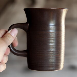 May include: A dark brown ceramic mug with a cylindrical body and a flared rim. The exterior surface features a series of horizontal, textured ridges that wrap around the middle section of the mug, providing a tactile grip and a matte finish.