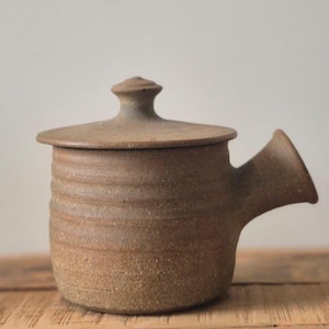 Peut inclure: Une théière en céramique marron avec un couvercle et un bec verseur. La théière a une surface texturée avec des rainures horizontales autour du corps. Le couvercle a un petit bouton sur le dessus. La théière est sur une surface en bois.