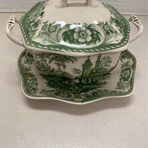 May include: Cream-colored soup tureen with a green floral and landscape design. The tureen has a lid with a handle and two side handles. It sits on a matching saucer with a scalloped edge. The design includes flowers, trees, and buildings.
