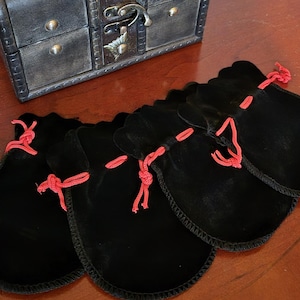 Pode incluir: Um baú decorativo em estilo antigo com acabamento metálico escuro, ferragens de latão e uma trava. Vários sacos de veludo preto com cordões vermelhos são dispostos em frente ao baú.
