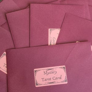 May include: A pile of burgundy envelopes, each with a pink label reading "Mystery Tarot Card." The envelopes are on a decorative silver tray, with a green and gold patterned surface in the background.