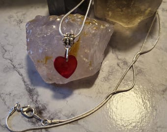 Op de afbeelding: Een zilveren ketting met een rode hartvormige hanger. De ketting ligt op een wit marmeren oppervlak met twee kristallen.