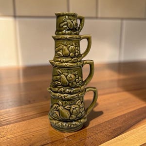 May include: A stack of four olive green ceramic measuring cups, each featuring a molded fruit design and a handle. The cups are labeled with measurements including 1/4 cup, 1/3 cup, 1/2 cup, and 1 cup, with each piece stacked vertically.