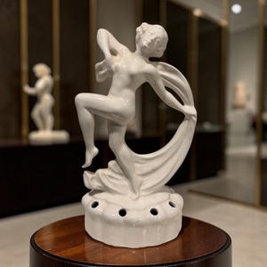 May include: A white ceramic figurine of a dancing figure with flowing fabric, mounted on a circular base featuring a series of small, evenly spaced holes around the perimeter. The figure is depicted in a dynamic pose with one leg raised.