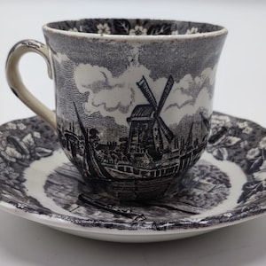 May include: A black and white teacup and saucer set with a windmill and cityscape design. The cup and saucer have a floral pattern around the rim.