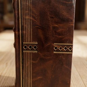 Puede incluir: Un libro encuadernado en cuero marrón con detalles dorados. La portada presenta líneas verticales doradas y patrones en forma de diamante. El libro está de pie sobre una superficie de madera, mostrando su cuero texturizado y diseño decorativo.