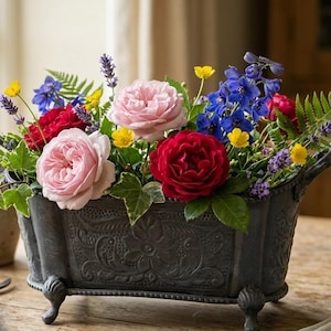 Pode incluir: Uma floreira retangular de metal cinza-escuro com padrões florais em relevo e quatro pés pequenos, preenchida com um arranjo vibrante de rosas cor-de-rosa e vermelhas, delfínios azuis, botões-de-ouro amarelos, alfazema e folhas de feto.