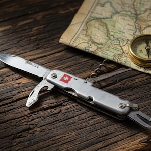 Puede incluir: Una navaja multiusos plateada con un emblema de la bandera suiza, una brújula y un mapa descansan sobre una superficie de madera envejecida. La navaja tiene una hoja, un abridor de botellas y otras herramientas. La brújula es dorada.