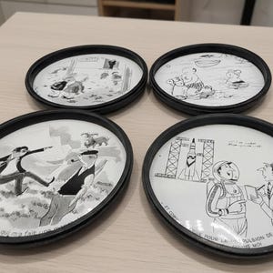 Puede incluir: Juego de cuatro posavasos con borde negro, cada uno con una ilustración de dibujos animados en blanco y negro. Las escenas muestran personas y animales en situaciones divertidas, con texto en francés. Un producto divertido y original.