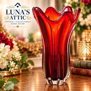 May include: A tall, vibrant red glass vase featuring a distinct wavy, scalloped rim and a vertical ribbed body. The base is clear glass, transitioning into a deep, translucent red hue that catches the light, creating a striking visual effect.