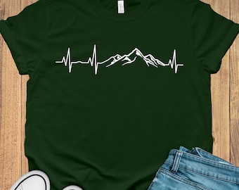Könnte beinhalten: Ein dunkelgrünes T-Shirt mit einer weißen Bergkette und einer Herzschlaglinie. Das Shirt liegt flach auf einer Holzoberfläche mit einem Paar weißen Sneakers und einer blauen Jeans.