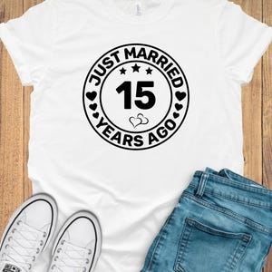 May include: White t-shirt with a black circular graphic that reads "Just Married 15 Years Ago." The graphic includes hearts, stars, and the number 15. The shirt is paired with blue jeans and white sneakers.