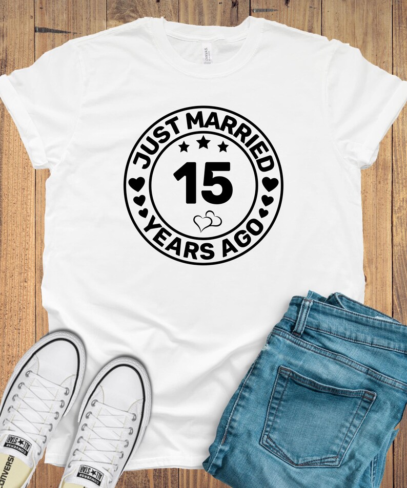 May include: White t-shirt with a black circular graphic that reads "Just Married 15 Years Ago." The graphic includes hearts, stars, and the number 15. The shirt is paired with blue jeans and white sneakers.