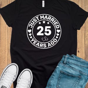 May include: Black t-shirt with a white circular graphic that reads "JUST MARRIED 25 YEARS AGO." The design includes stars, hearts, and the number 25. The image also shows white sneakers and blue jeans.