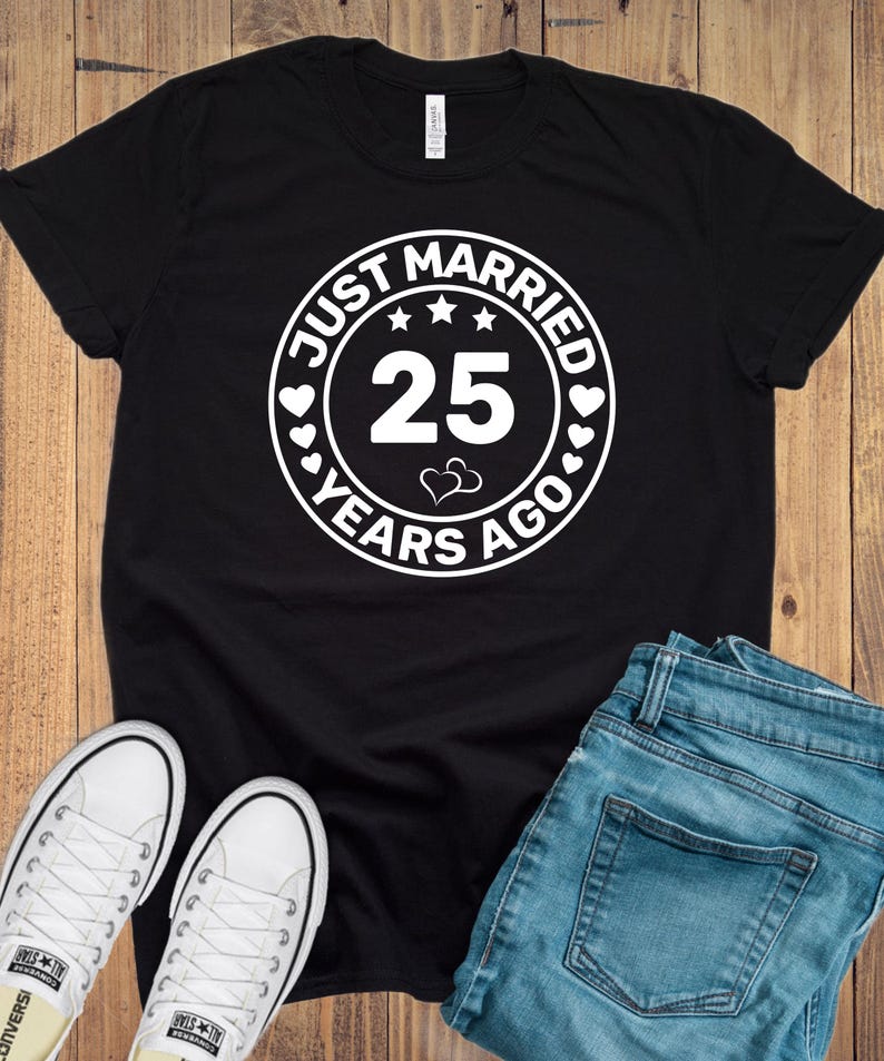 May include: Black t-shirt with a white circular graphic that reads "JUST MARRIED 25 YEARS AGO." The design includes stars, hearts, and the number 25. The image also shows white sneakers and blue jeans.