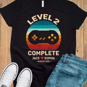 Puede incluir: Camiseta negra con un gr&aacute;fico de mando de videojuego de estilo retro y el texto "Level 2 Complete Jack &hearts; Sophia - August 2021 - "
