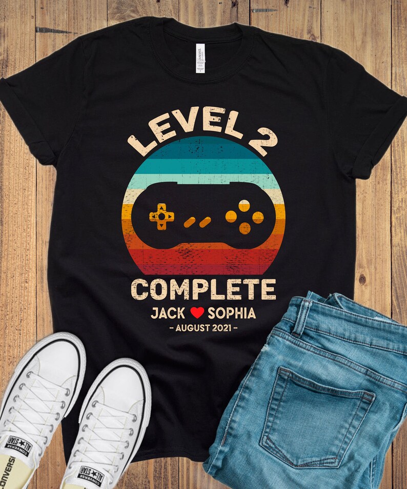 Puede incluir: Camiseta negra con un gr&aacute;fico de mando de videojuego de estilo retro y el texto "Level 2 Complete Jack &hearts; Sophia - August 2021 - "