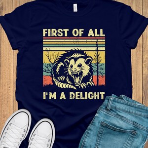 May include: A navy blue t-shirt with a vintage-style graphic of a possum with the text "First of all I'm a delight".