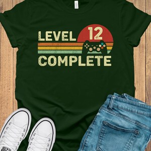 May include: Dark green t-shirt with the text "LEVEL 12 COMPLETE" in a retro font, a red circle with the number 12, and a game controller graphic. The shirt is paired with blue jeans and white sneakers.