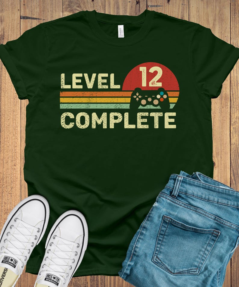 May include: Dark green t-shirt with the text "LEVEL 12 COMPLETE" in a retro font, a red circle with the number 12, and a game controller graphic. The shirt is paired with blue jeans and white sneakers.