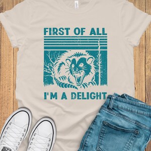 May include: A beige t-shirt with a vintage-style graphic of a raccoon with the text "First of all I'm a delight".