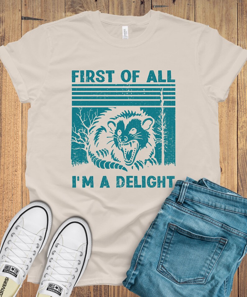 May include: A beige t-shirt with a vintage-style graphic of a raccoon with the text "First of all I'm a delight".