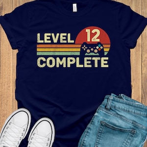 May include: Navy blue t-shirt with the text "LEVEL 12 COMPLETE" in a retro design. The design includes a red circle, a game controller, and horizontal stripes in yellow, orange, and green. White sneakers and blue jeans are also visible.