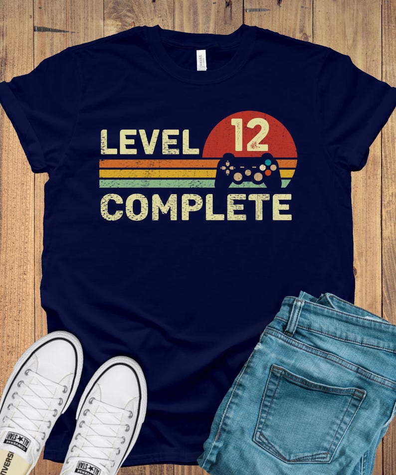 May include: Navy blue t-shirt with the text "LEVEL 12 COMPLETE" in a retro design. The design includes a red circle, a game controller, and horizontal stripes in yellow, orange, and green. White sneakers and blue jeans are also visible.