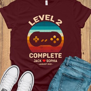 Puede incluir: Una camiseta color granate con un gr&aacute;fico de mando de videojuego de estilo retro. El texto "Level 2 Complete" est&aacute; por encima del mando, y el texto "Jack Sophia - August 2021 -" est&aacute; por debajo del mando.
