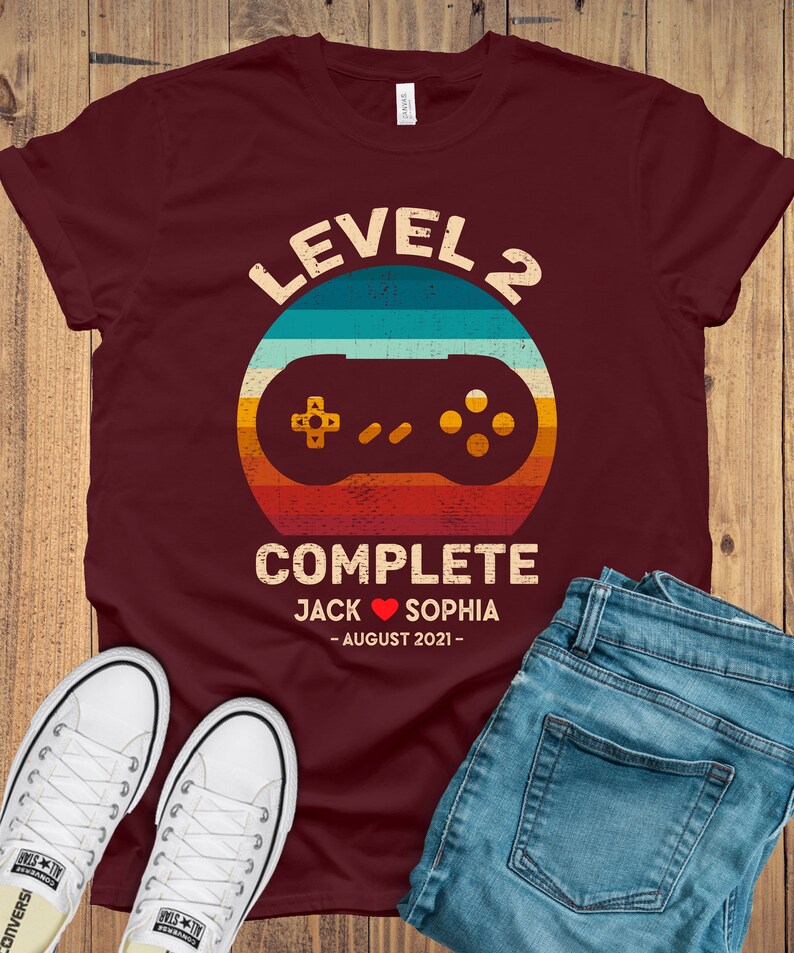 Puede incluir: Una camiseta color granate con un gr&aacute;fico de mando de videojuego de estilo retro. El texto "Level 2 Complete" est&aacute; por encima del mando, y el texto "Jack Sophia - August 2021 -" est&aacute; por debajo del mando.