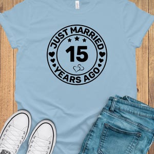 May include: A light blue t-shirt with the text "JUST MARRIED 15 YEARS AGO" in a black circular design. The shirt is paired with blue jeans and white sneakers.