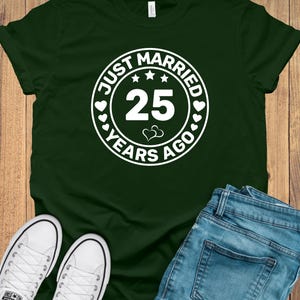 May include: Dark green t-shirt with a white circular graphic that reads "JUST MARRIED 25 YEARS AGO". The image also shows blue jeans and white canvas sneakers, suggesting a casual style.