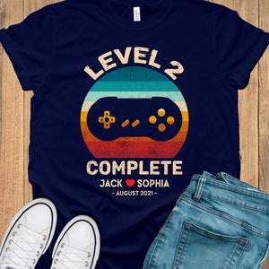 Puede incluir: Camiseta azul marino con un gr&aacute;fico de mando de videojuego de estilo retro. El texto "Level 2 Complete" est&aacute; por encima del mando, y el texto "Jack Sophia - August 2021 -" est&aacute; por debajo del mando.