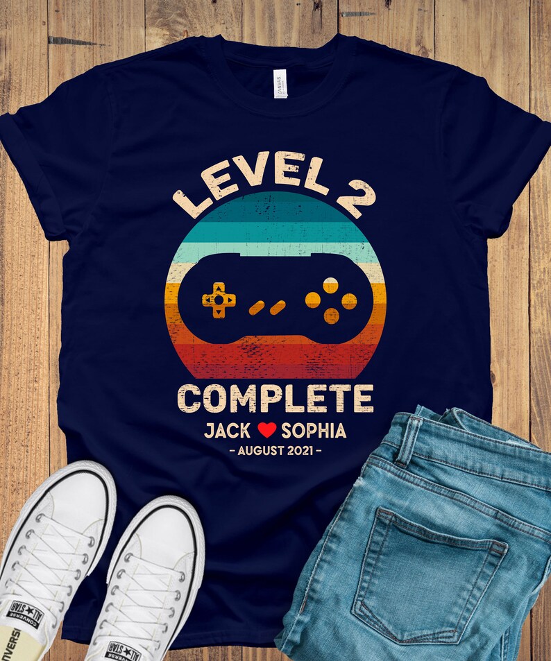 Puede incluir: Camiseta azul marino con un gr&aacute;fico de mando de videojuego de estilo retro. El texto "Level 2 Complete" est&aacute; por encima del mando, y el texto "Jack Sophia - August 2021 -" est&aacute; por debajo del mando.