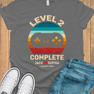 Puede incluir: Camiseta gris con un gr&aacute;fico de mando de videojuego de estilo retro y el texto "Level 2 Complete Jack &hearts; Sophia - August 2021 - "