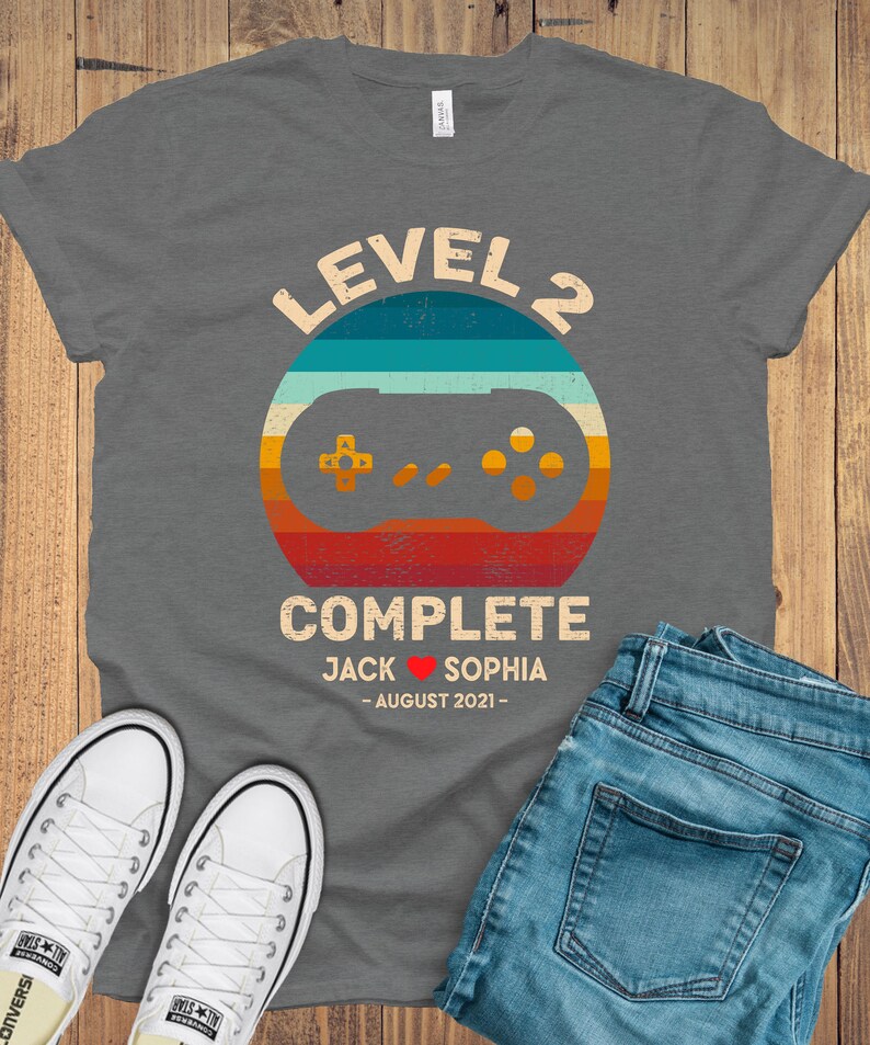 Puede incluir: Camiseta gris con un gr&aacute;fico de mando de videojuego de estilo retro y el texto "Level 2 Complete Jack &hearts; Sophia - August 2021 - "