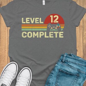 May include: A gray t-shirt with the text "LEVEL 12 COMPLETE" in a retro font. The design includes a red circle with the number 12, a controller graphic, and colorful stripes. White sneakers and blue jeans are also visible.